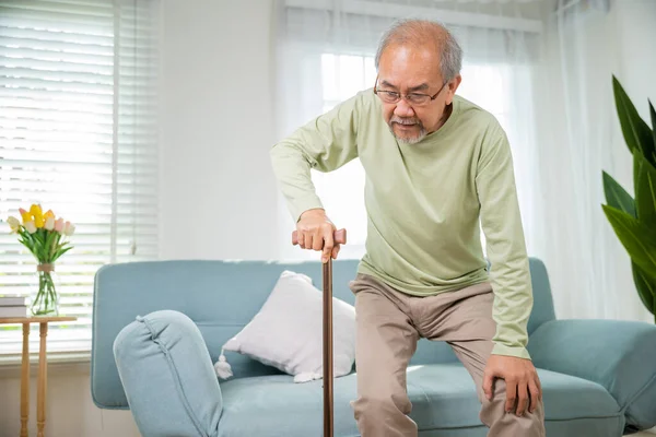 メガネタイプのアジアの高齢者は 杖スティックを歩くと自宅で歩くとソファから立ち上がるには 膝痛の痛みを抱えている高齢者杖のハンドル 退職医療 — ストック写真