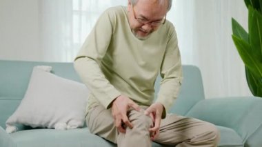 Kanepe oturma odasında diz eklem ağrısı çeken yaşlı bir adam, evde kemik ağrısı, yaşlı bir adam dizinde ağrı, acı verici, tendon ameliyatından sonra dizinde ağrısı olan mutsuz yaşlı bir adam.