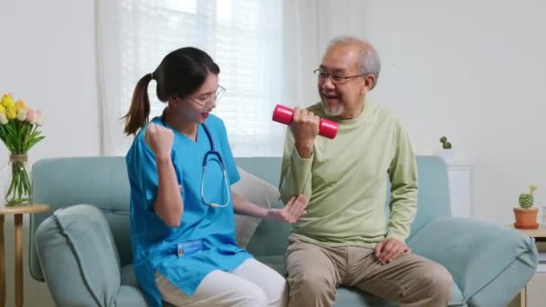 アジアの看護師理学療法士は 老人ホームでダンベルを持ち上げるのに役立ちます 若い看護師はダンベル運動を使用してソファに座って高齢者のケアサポートトレーニングを取ります — ストック動画