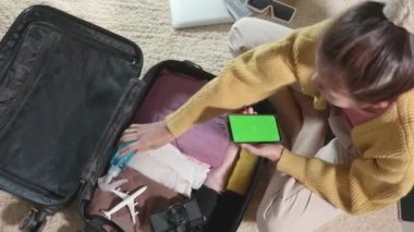 Woman using smartphone blank green screen for checklist and packing suitcase bag for travel trip weekend vacation at home, female check note purveyance or baggage on mobile phone