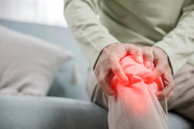 Kanepe oturma odasında diz eklem ağrısı çeken yaşlı bir adam, evde kemik ağrısı, yaşlı bir adam dizinde ağrı, acı verici, tendon ameliyatından sonra dizinde ağrısı olan mutsuz yaşlı bir adam.