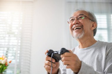 Asyalı son sınıf öğrencisi evde video oyunu oynamaktan zevk alıyor, olgun erkek elleri oyun kumandası kullanıyor, emeklilik gülümsemesinde komik, kanepede oturmuş oyun oynuyor.