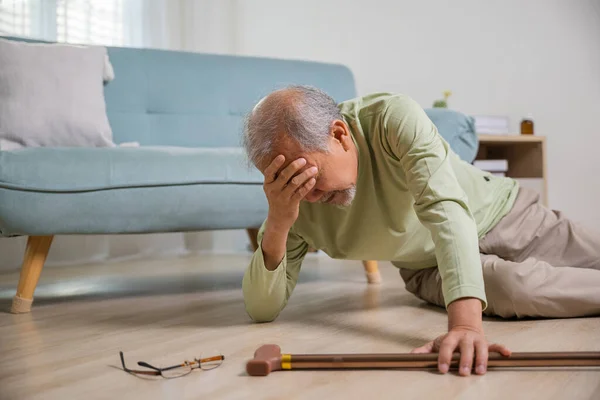 Asian senior man falling down lying on floor at home alone with wooden walking stick in living room, Elderly man headache after fall down, Health care and medicine concept