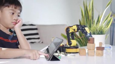 Asian little kid boy using digital tablet computer on robotic arm machine in workshop, Child learning getting lesson programming control robot arm pick up wood block, Technology science education