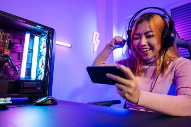 Winning. Happy Gamer playing video game online with smartphone with neon lights she raises hands to wins celebrating, confused Asian woman wearing gaming headphones feeling excited at home
