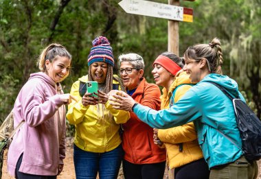 Group of women with different ages and ethnicities having fun using mobile smartphone while walking in foggy forest - Adventure and travel people concept clipart