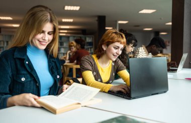 Kütüphanede dizüstü bilgisayar kullanan ve kitap okuyan genç üniversite öğrencileri - Okul eğitimi kavramı