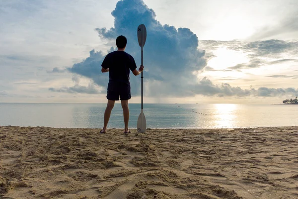 澄んだ砂浜に立つ男と朝の日の出を背景にパドルを保持 — ストック写真