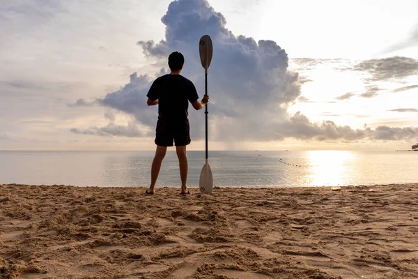 澄んだ砂浜に立つ男と朝の日の出を背景にパドルを保持 — ストック写真
