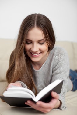 Happy young woman reading book on sofa clipart