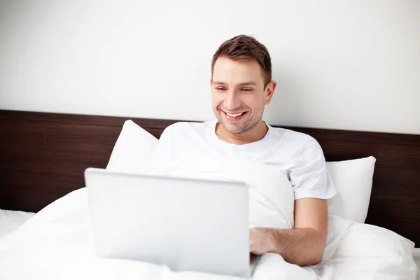 Χαρούμενος νεαρός άνδρας κάθεται στο κρεβάτι με το lap-top — Φωτογραφία Αρχείου
