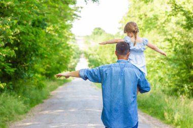 Omuzlarında ebeveyni olan mutlu çocuk park arka planında yol boyunca yürür.