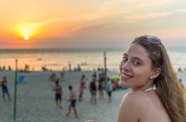 Genç bir kadın gün batımında kumsalda kameraya bakıyor.