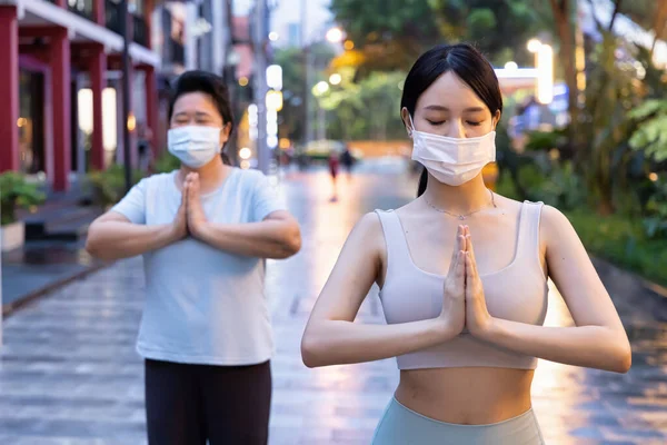 Personal Yoga trainer with face mask helping senior woman doing yoga meditation