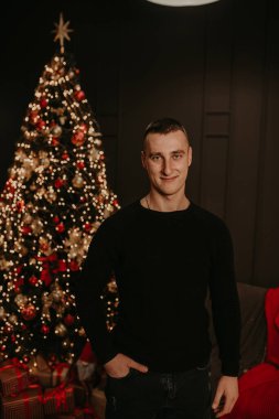 Siyah elbiseli yakışıklı genç adam çelenklerle bir Noel ağacının yanında. Yeni yıl için dekore edilmiş bir ev. Noel sabahı. Dairenin içi. Sevgililer Günü kutlaması