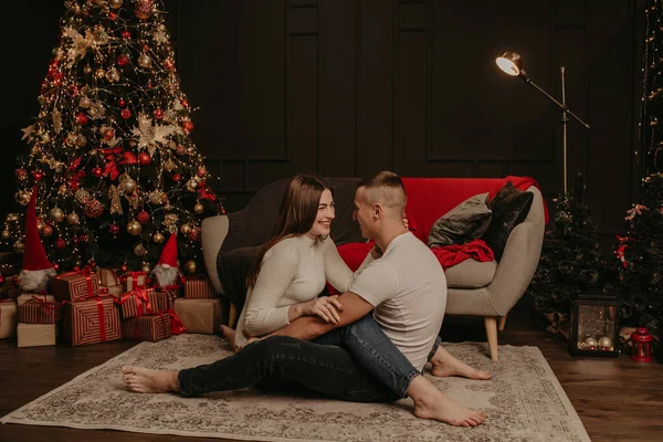 Verliebtes Paar Mann und Frau sitzen im Schneidersitz auf dem Boden und umarmen und küssen Weihnachtsbaum. — Stockfoto