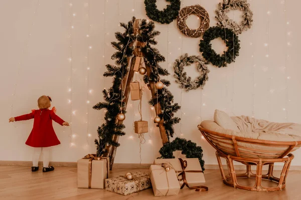 Çocuk beyaz duvarda çelenklerle duruyor Noel ağacında paketlenmiş hediyelerle