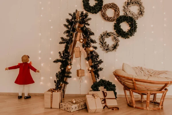 Παιδί στέκεται σε λευκό τοίχο με γιρλάντες Χριστουγεννιάτικο δέντρο με τυλιγμένα δώρα Royalty Free Εικόνες Αρχείου