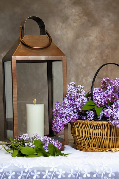 迷人的形象与一篮子新鲜切紫丁香花与铜大灯笼旁边的蜡烛 背景是一个纹理混合的棕色 他们都坐在精致的花边桌布上 — 图库照片