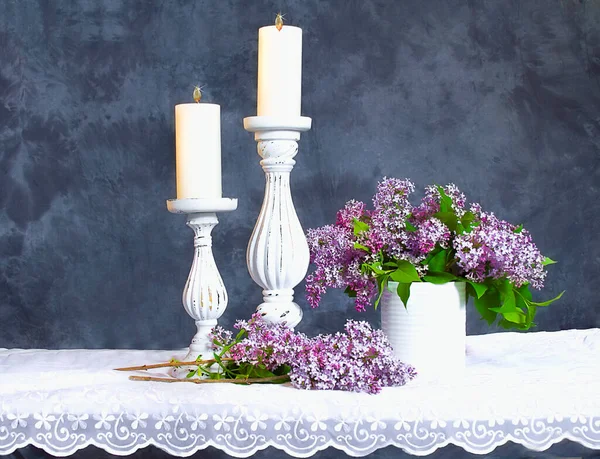新鲜的丁香花插在一个白色陶瓷花瓶里 里面有两个柱子烛台和蜡烛 背景是蓝色纹理状的材料 桌面是柔软的白色花边 — 图库照片