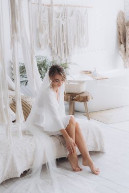 Gelinin kırsal tarzda bir sabah, beyaz bornoz ve beyaz bir buket içinde yatağın ve banyonun yanında bir fotoğraf stüdyosunun, bir otelin penceresinin yanında