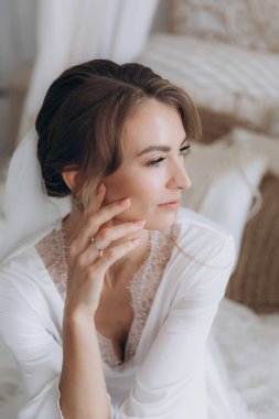 Gelinin kırsal tarzda bir sabah, beyaz bornoz ve beyaz bir buket içinde yatağın ve banyonun yanında bir fotoğraf stüdyosunun, bir otelin penceresinin yanında