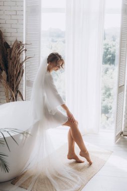 Gelinin kırsal tarzda bir sabah, beyaz bornoz ve beyaz bir buket içinde yatağın ve banyonun yanında bir fotoğraf stüdyosunun, bir otelin penceresinin yanında