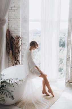 Gelinin kırsal tarzda bir sabah, beyaz bornoz ve beyaz bir buket içinde yatağın ve banyonun yanında bir fotoğraf stüdyosunun, bir otelin penceresinin yanında