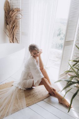 Gelinin kırsal tarzda bir sabah, beyaz bornoz ve beyaz bir buket içinde yatağın ve banyonun yanında bir fotoğraf stüdyosunun, bir otelin penceresinin yanında