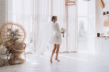 Gelinin kırsal tarzda bir sabah, beyaz bornoz ve beyaz bir buket içinde yatağın ve banyonun yanında bir fotoğraf stüdyosunun, bir otelin penceresinin yanında