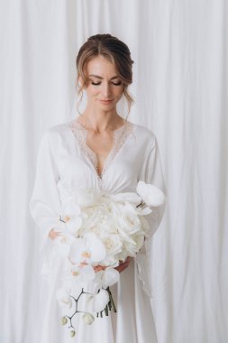 Gelinin kırsal tarzda bir sabah, beyaz bornoz ve beyaz bir buket içinde yatağın ve banyonun yanında bir fotoğraf stüdyosunun, bir otelin penceresinin yanında