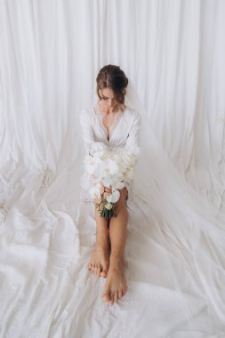 Gelinin kırsal tarzda bir sabah, beyaz bornoz ve beyaz bir buket içinde yatağın ve banyonun yanında bir fotoğraf stüdyosunun, bir otelin penceresinin yanında