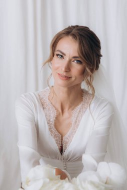 Gelinin kırsal tarzda bir sabah, beyaz bornoz ve beyaz bir buket içinde yatağın ve banyonun yanında bir fotoğraf stüdyosunun, bir otelin penceresinin yanında