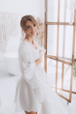 Gelinin kırsal tarzda bir sabah, beyaz bornoz ve beyaz bir buket içinde yatağın ve banyonun yanında bir fotoğraf stüdyosunun, bir otelin penceresinin yanında