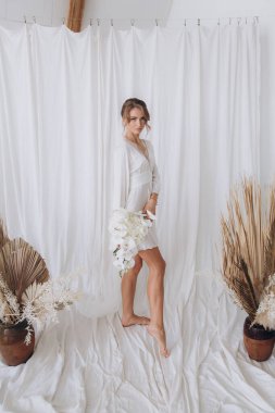 Gelinin kırsal tarzda bir sabah, beyaz bornoz ve beyaz bir buket içinde yatağın ve banyonun yanında bir fotoğraf stüdyosunun, bir otelin penceresinin yanında