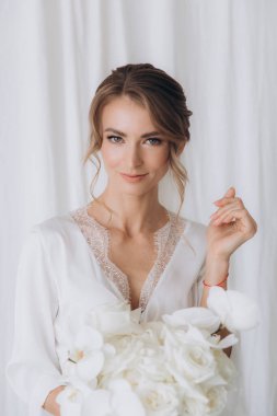 Gelinin kırsal tarzda bir sabah, beyaz bornoz ve beyaz bir buket içinde yatağın ve banyonun yanında bir fotoğraf stüdyosunun, bir otelin penceresinin yanında