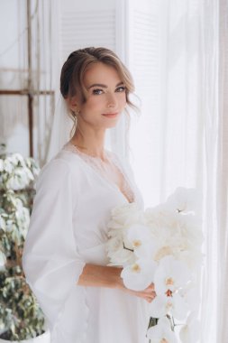 Gelinin kırsal tarzda bir sabah, beyaz bornoz ve beyaz bir buket içinde yatağın ve banyonun yanında bir fotoğraf stüdyosunun, bir otelin penceresinin yanında