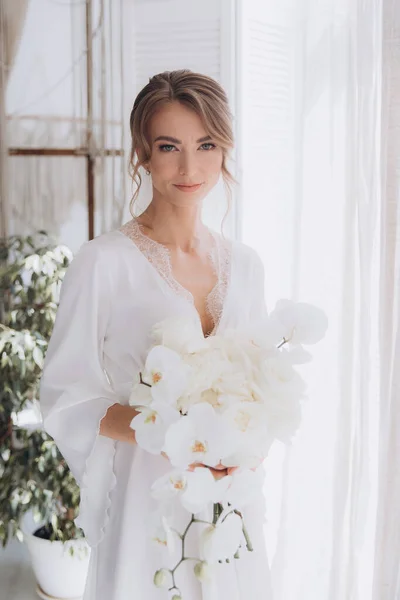 Gelinin kırsal tarzda bir sabah, beyaz bornoz ve beyaz bir buket içinde yatağın ve banyonun yanında bir fotoğraf stüdyosunun, bir otelin penceresinin yanında