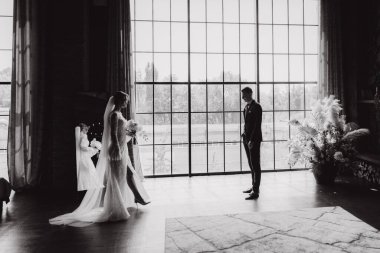 Siyah takım elbiseli damadın ilk buluşması ve siyah arka planda bir fotoğraf stüdyosunun, otelin içinde beyaz gelinlik içinde bir buket.