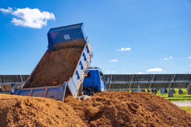 A blue six-wheeled vehicle is pouring orange soil onto the floor of an industrial site to fill in a well damaged by erosion water at the solar power plant.