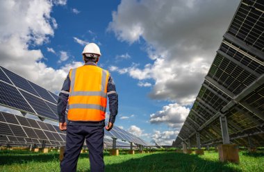 Technician dressed in blue uniform and in an orange reflective vest and wearing a safety helmet stands facing a solar panel at solar power plant, Photovoltaic module idea for clean energy production.