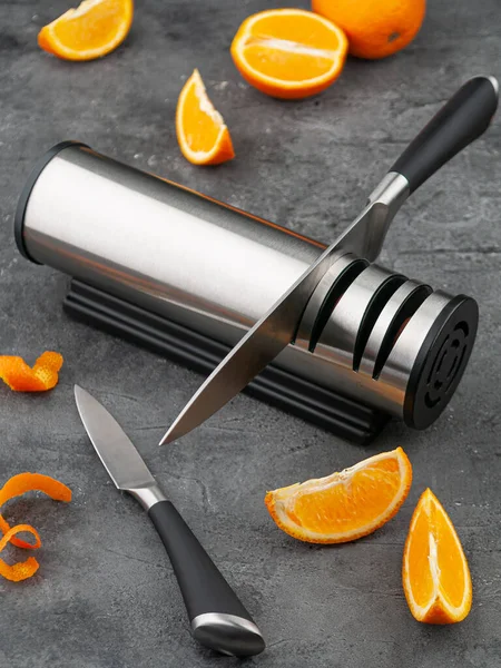 Electric knife sharpener. Cylinder-shaped made of metal and plastic. A gadget for the kitchen and interior. Gray concrete background. Nearby are kitchen knives and sliced oranges.