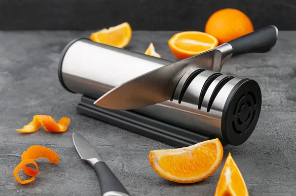 Electric knife sharpener. Cylinder-shaped made of metal and plastic. A gadget for the kitchen and interior. Gray concrete background. Nearby are kitchen knives and sliced oranges.