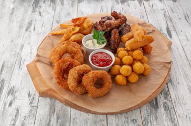 Bira için atıştırmalık bir şeyler. Mangal kanatları, kroketler, peynir çubukları, soğan halkaları ve cips. Domates ve peynir soslu. Yuvarlak ahşap bir tahtada. Hafif ahşap zemin. Yukarıdan görüntüle.