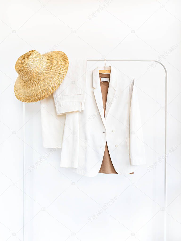 Womens jacket, pants and straw hat in shades of milky white with copy space. Rack with stylish female clothes on wooden hangers next to white wall. Clothing retails concept. Advertise, sale, fashion.