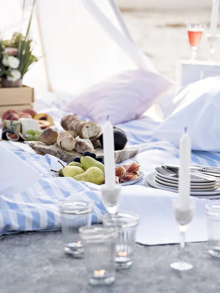 Various delicious food and beverages for romantic picnic lying amidst pillows in light tent. Summer brunch or dinner organised on a sea shore for friends. Elegant place for romantic date. Glamping.