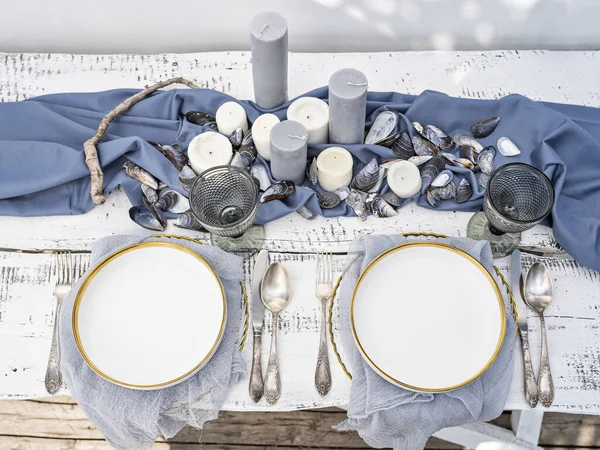 Sunny luxury table setting in marine style with candles, mussels shells and floating wood. Table served for engagement, wedding, romantic dinner or festive event. Outdoor glamorous catering or dining.