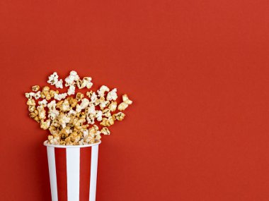 Patlamış mısır parlak kırmızı arka planda beyaz ve kırmızı çizgili bir kovadan döküldü. Sinema, sinema, film gecesi ve eğlence konsepti. Üstten, üstten, üstten.