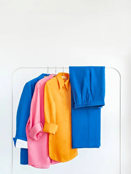 Woman\'s jacket, pants and linen shirts in bright colours with space for text. Rack with stylish female clothes on wooden hangers next to white wall. Clothing retails concept. Advertise, sale, fashion.