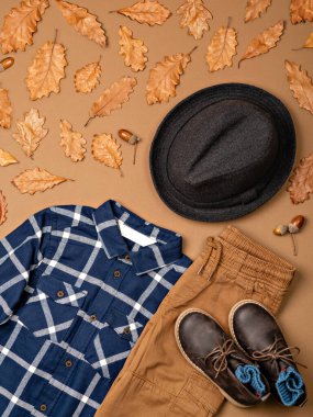 Küçük çocuğun kompozisyonu sıcak kıyafetler, ayakkabılar ve kahverengi arka planda yün bir şapka. Sonbahar için meşe yapraklı çocuk kıyafeti. Çocuk modası. Kış, sonbahar koleksiyonu. Organik pamuk. 